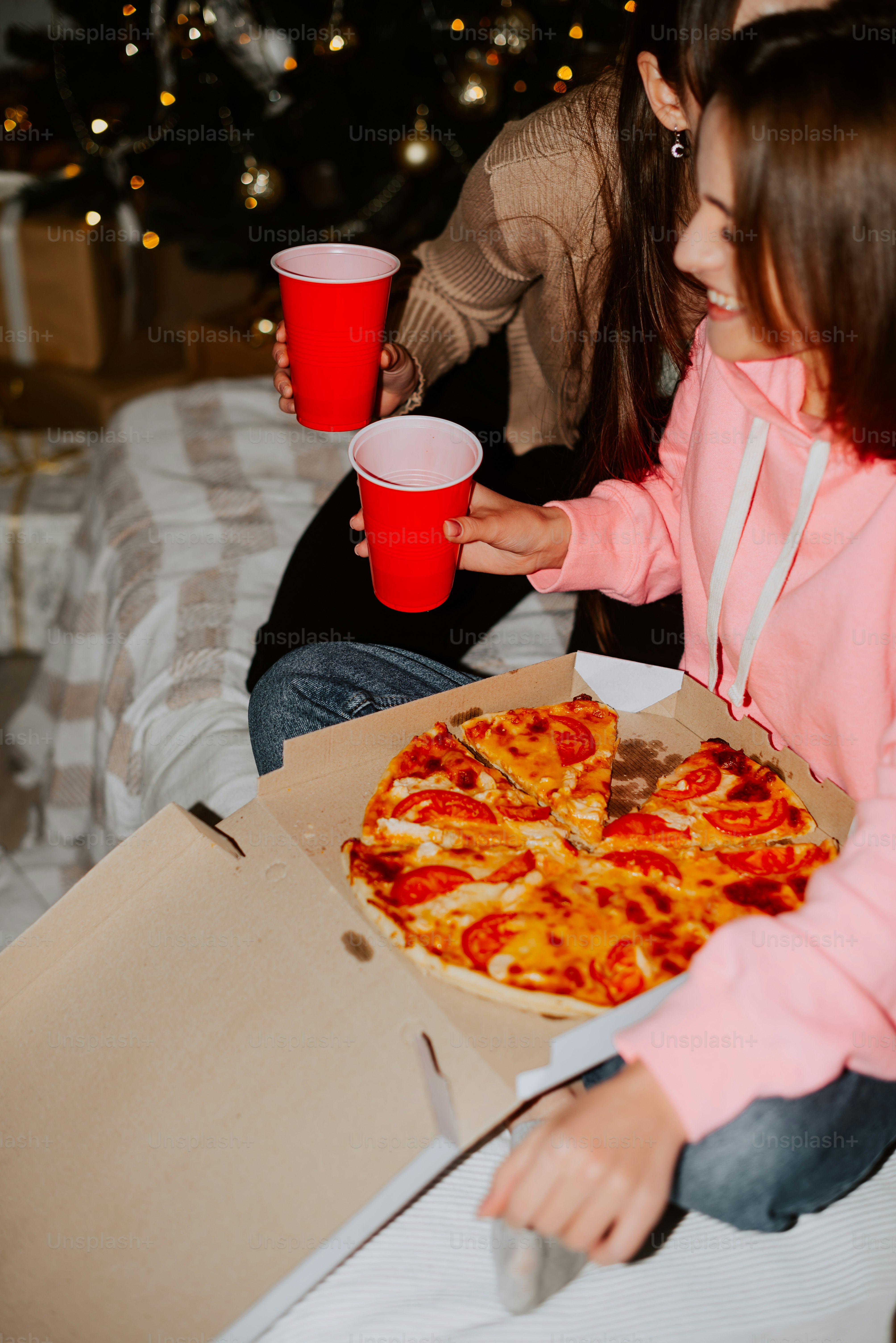 two girls sitting on a bed eating pizza and drinking red cups