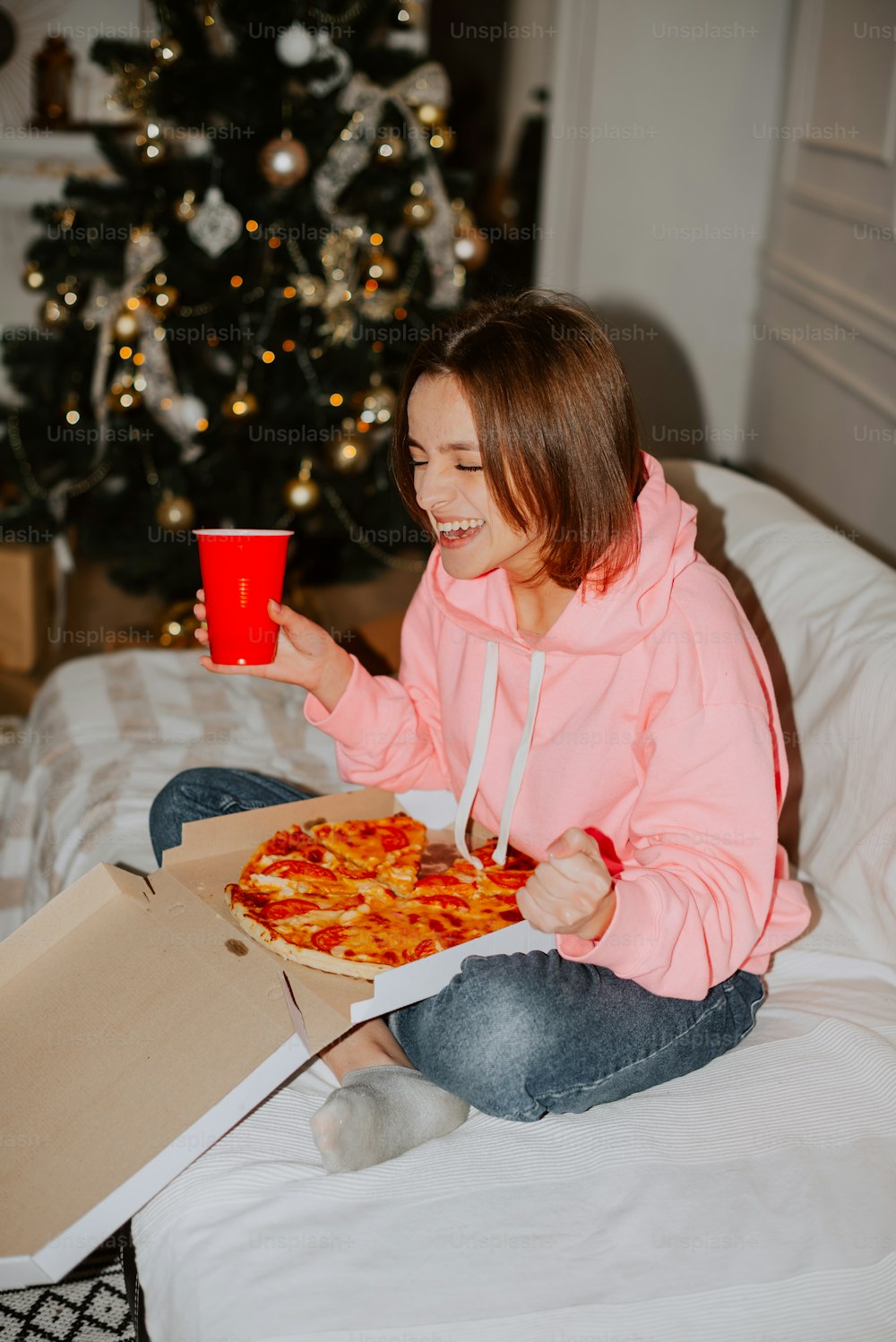 Una mujer sentada en una cama sosteniendo una taza roja y una pizza