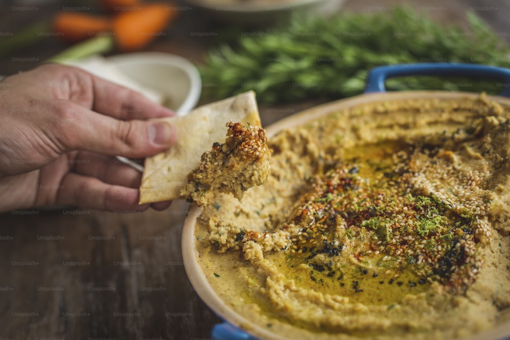 a hand holding a piece of pita bread over a bowl of hummus