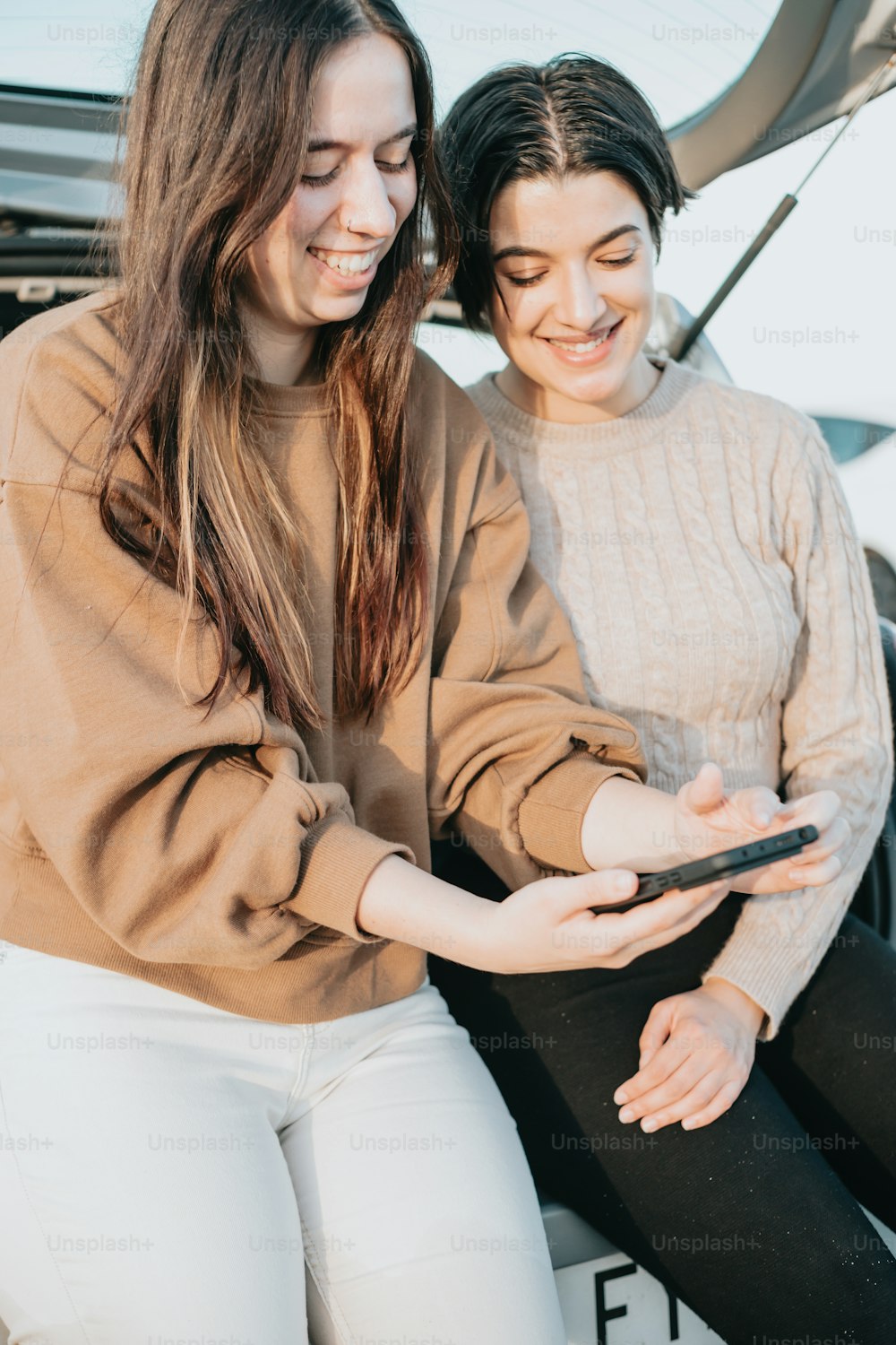 two women sitting on the hood of a car looking at a cell phone