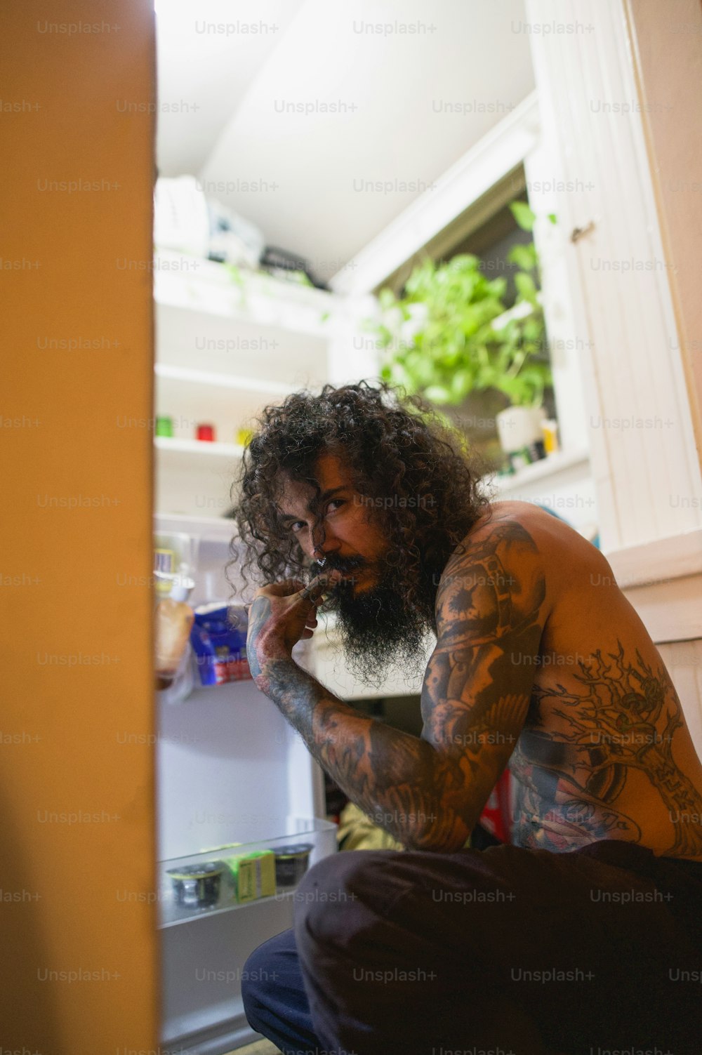 a man with long hair and tattoos sitting in front of a refrigerator