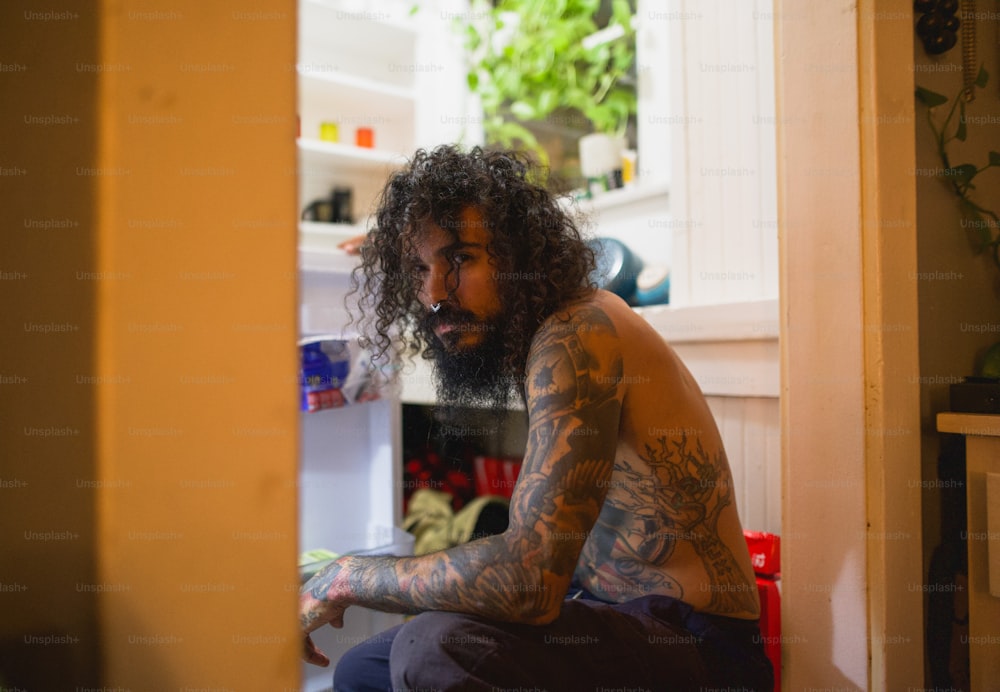 a man with long hair and tattoos sitting in front of a refrigerator