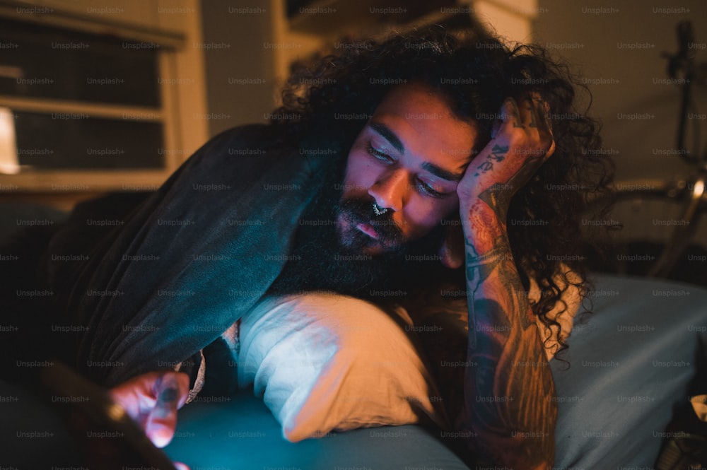 a man with long hair laying on a bed looking at a cell phone