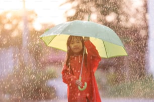 Asiatische Kinder breiten Regenschirme aus und spielen im Regen, sie trägt Regenkleidung.