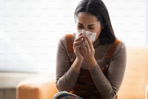 Asian woman sneezes. She uses a tissue to cover her mouth and she is working at home.