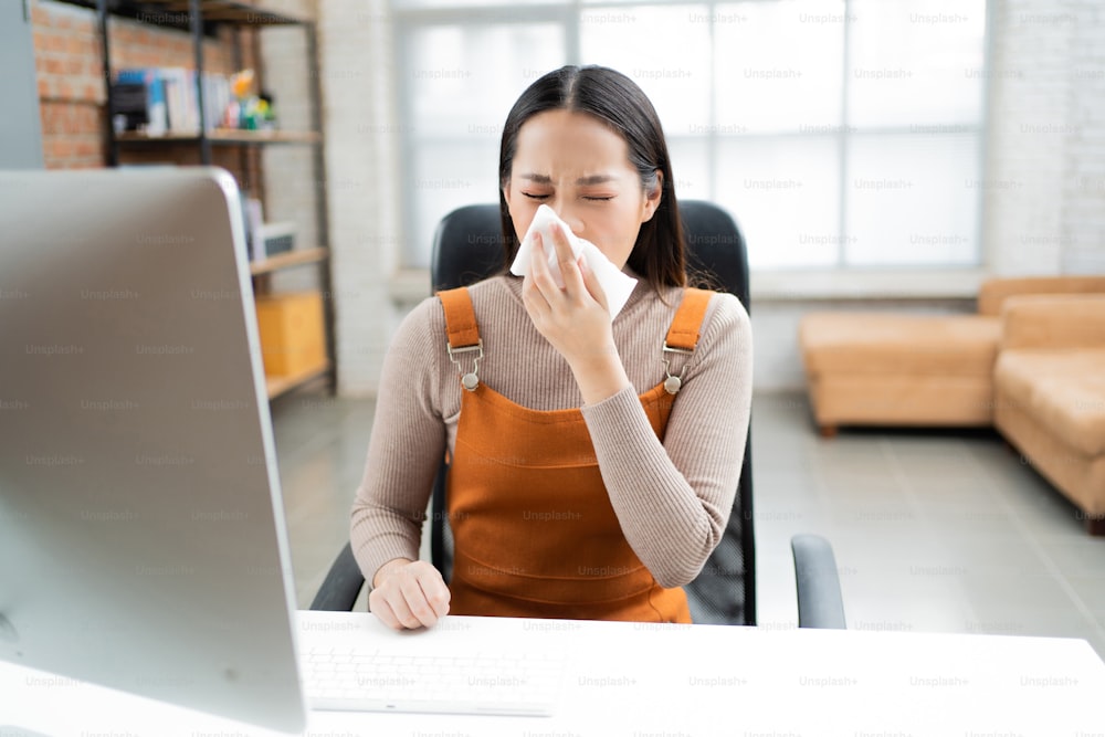 Asian woman sneezes. She uses a tissue to cover her mouth and she is working at home.
