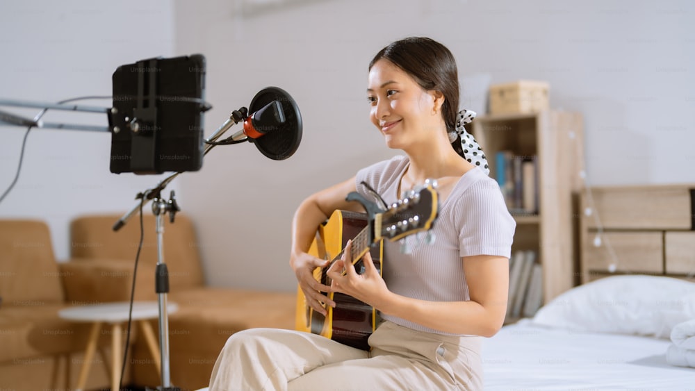 Asiatische Frau, die in ihrem Schlafzimmer Gitarre spielt und Gesang aufnimmt. Sie ist live.