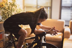 Cycliste asiatique. Elle fait de l’exercice à la maison. En faisant du vélo sur l’entraîneur et en jouant à des jeux de vélo en ligne