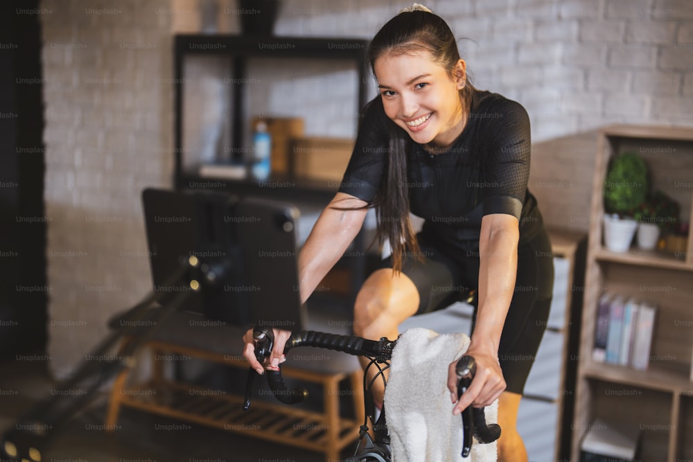 아시아 여자 사이클리스트. 그녀는 집에서 운동하고 있습니다. 트레이너에서 자전거를 타고 온라인 자전거 게임을 플레이하여