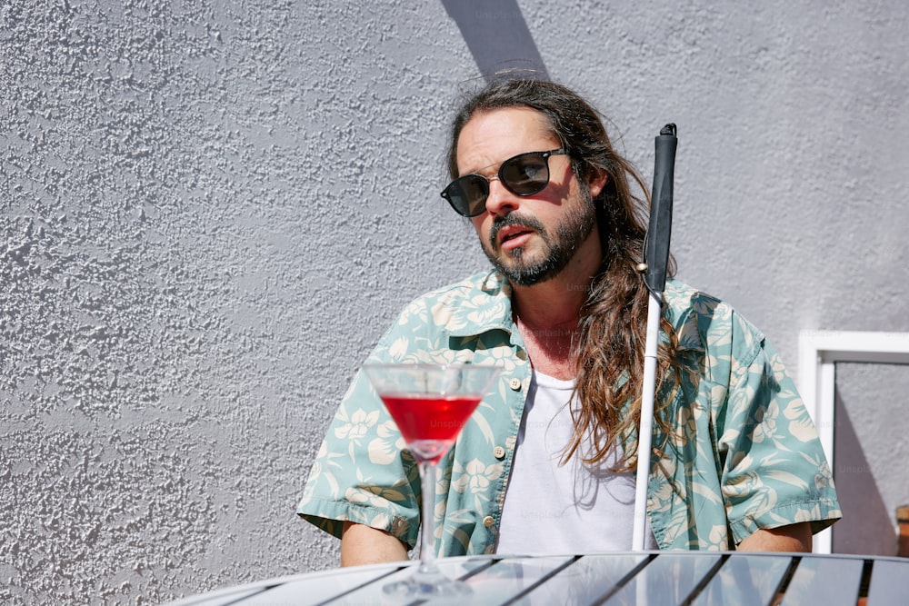 Un homme aux cheveux longs et aux lunettes de soleil tenant un verre