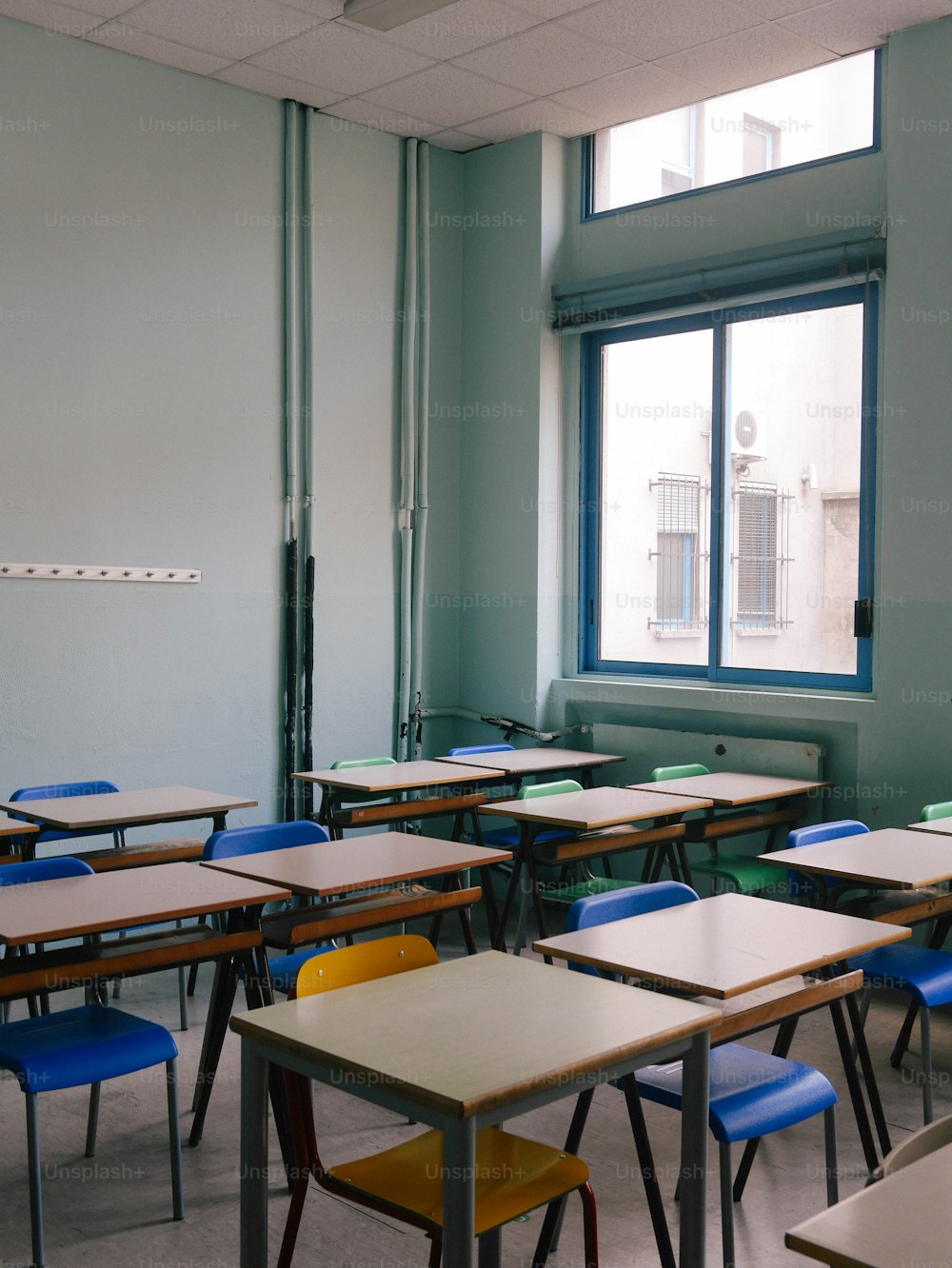 uma sala de aula cheia de carteiras e cadeiras ao lado de uma janela