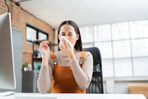 La donna asiatica starnutisce. Usa un fazzoletto per coprirsi la bocca e sta lavorando a casa.