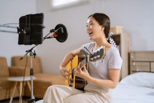 Asiatische Frau, die in ihrem Schlafzimmer Gitarre spielt und Gesang aufnimmt. Sie ist live.