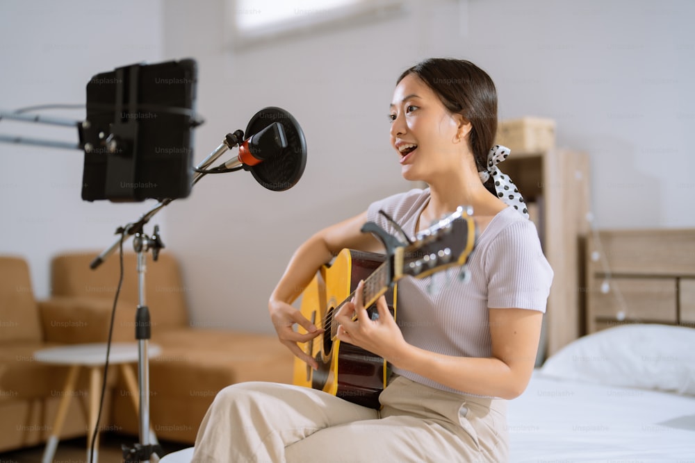Asian woman playing guitar and recording vocals in her bedroom. She's live.