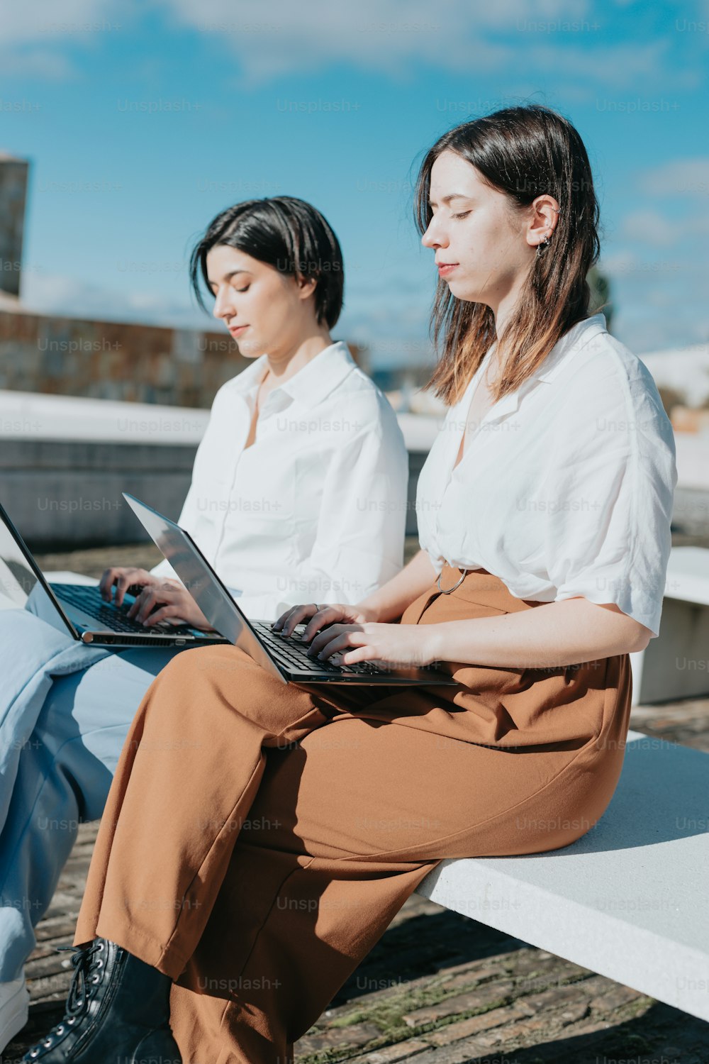 two women sitting on a bench with their laptops
