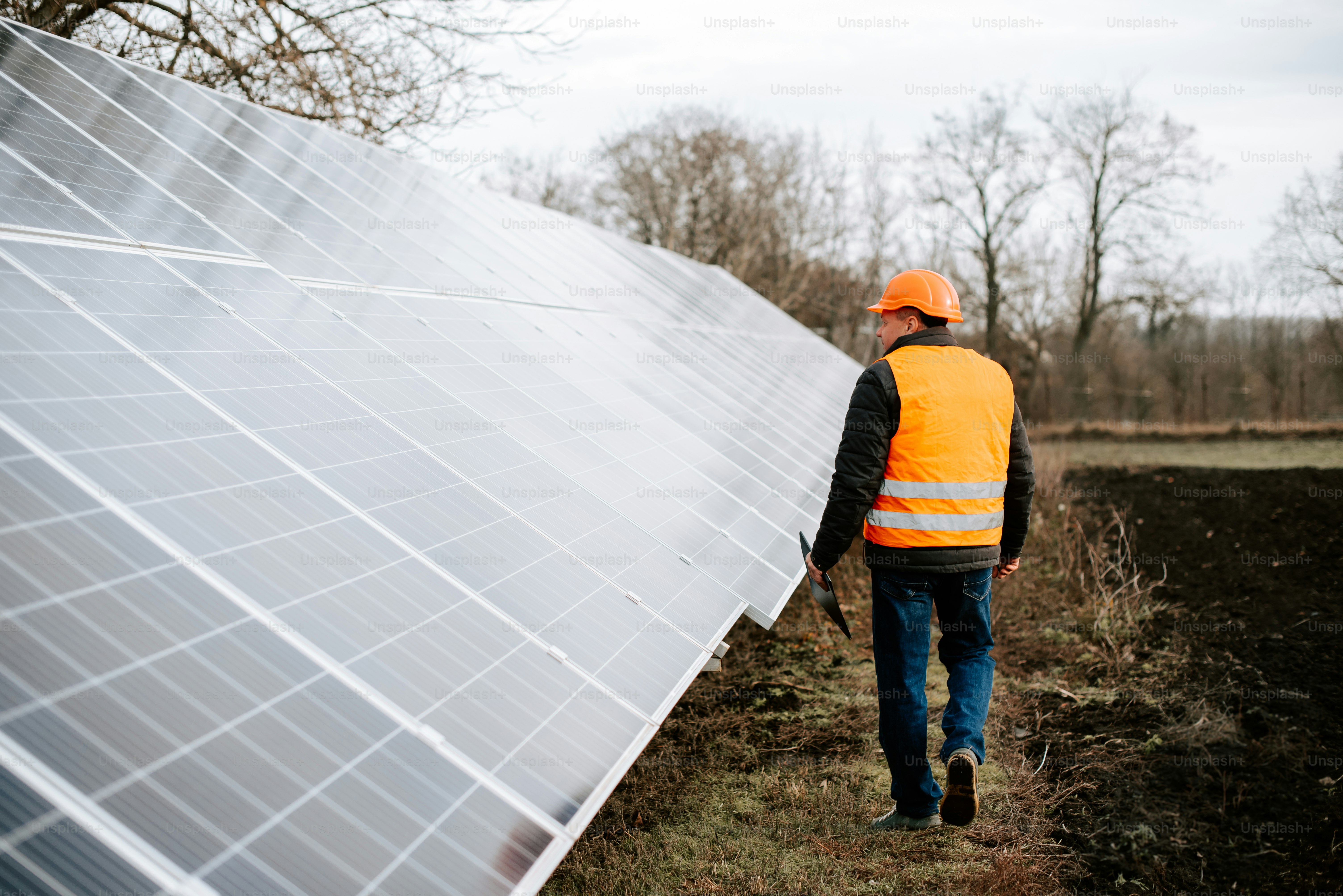 Ein Mann in orangefarbener Weste und Schutzhelm geht neben einem Solarpanel