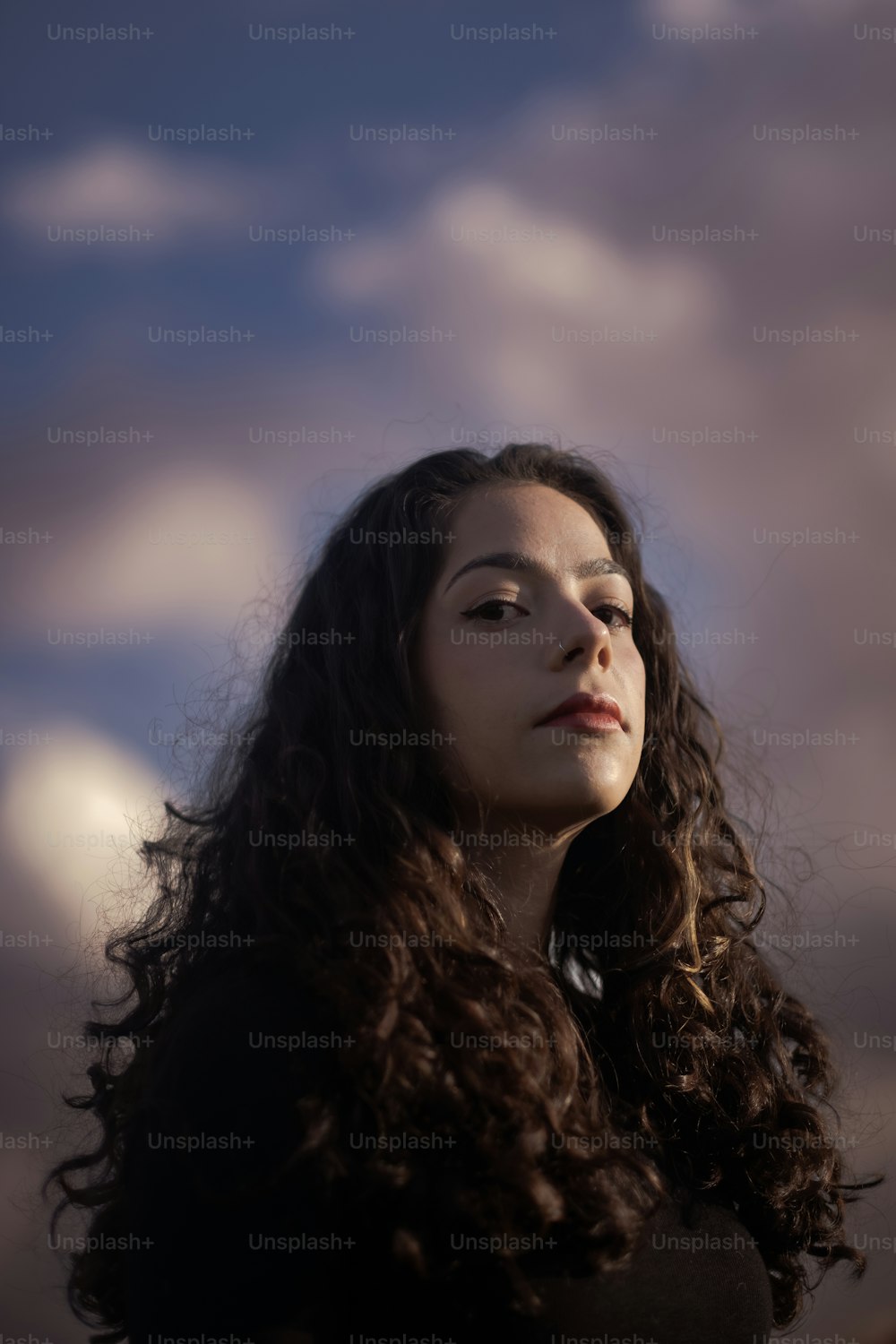 a woman with long curly hair standing in front of a cloudy sky