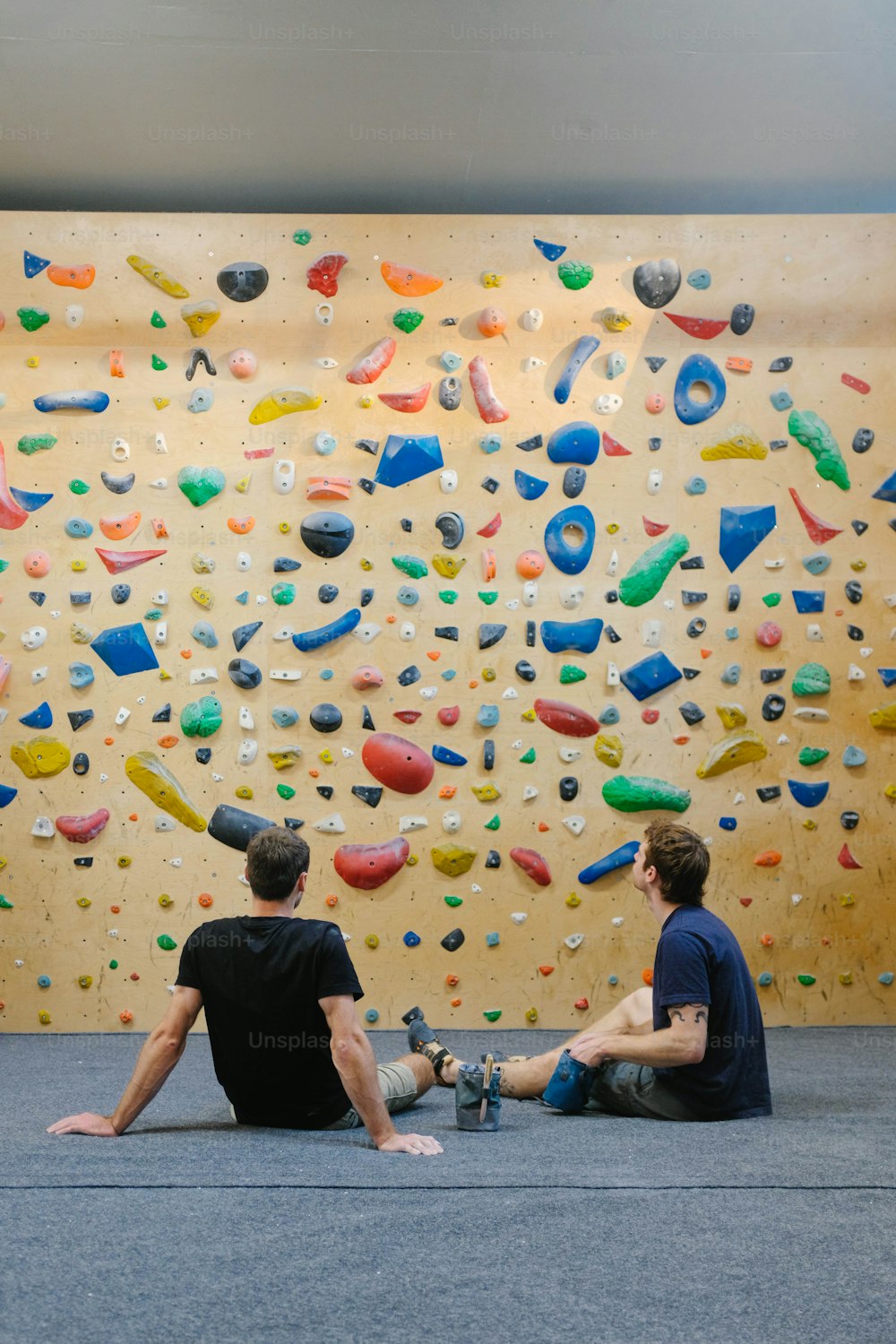 two men sitting on the floor in front of a climbing wall