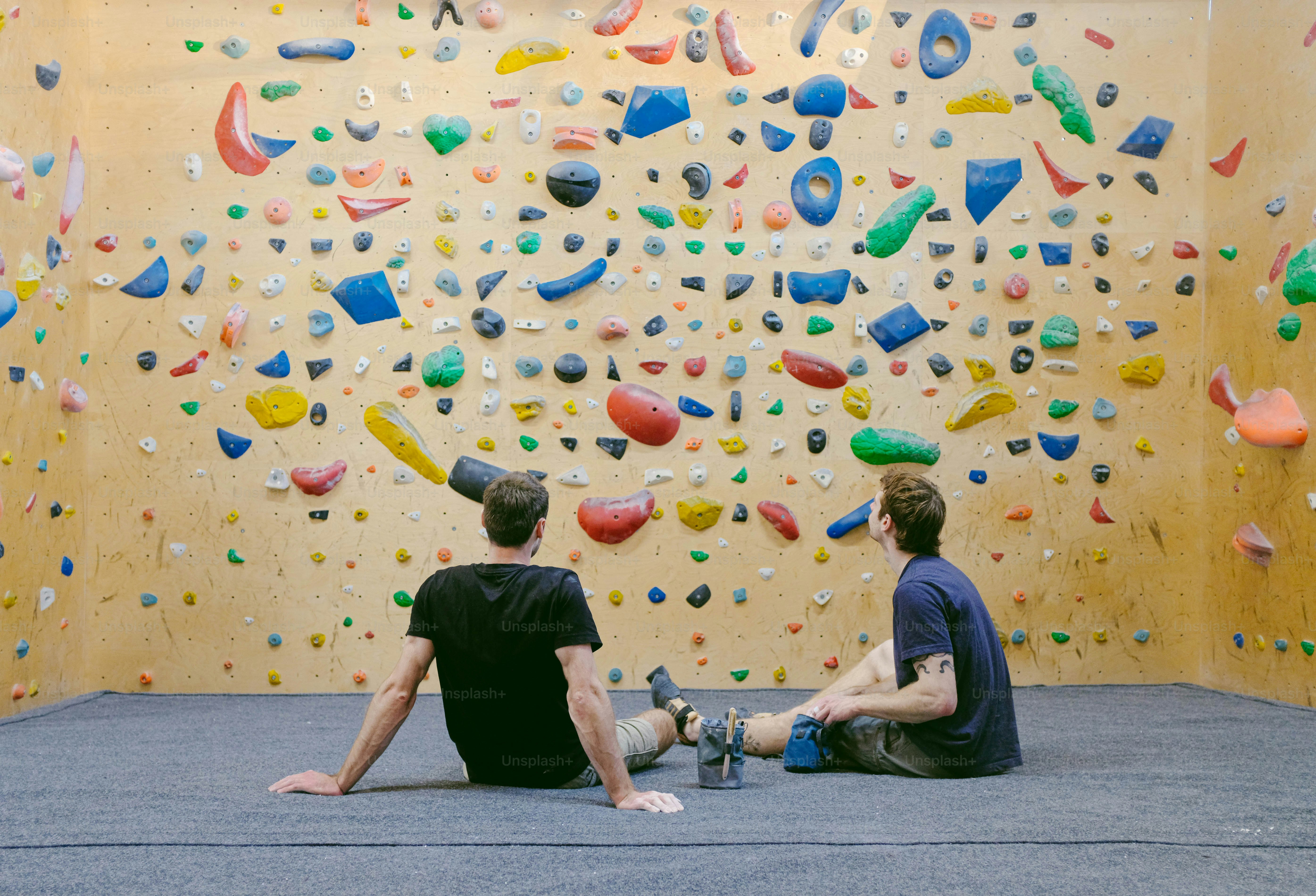 two men sitting on the floor in front of a climbing wall