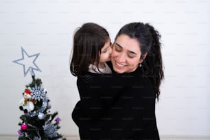 a woman kissing a little girl next to a christmas tree