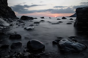 the sun is setting over the ocean with rocks in the foreground