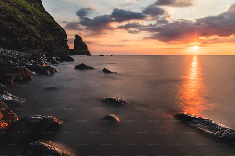 the sun is setting over the ocean with rocks in the foreground