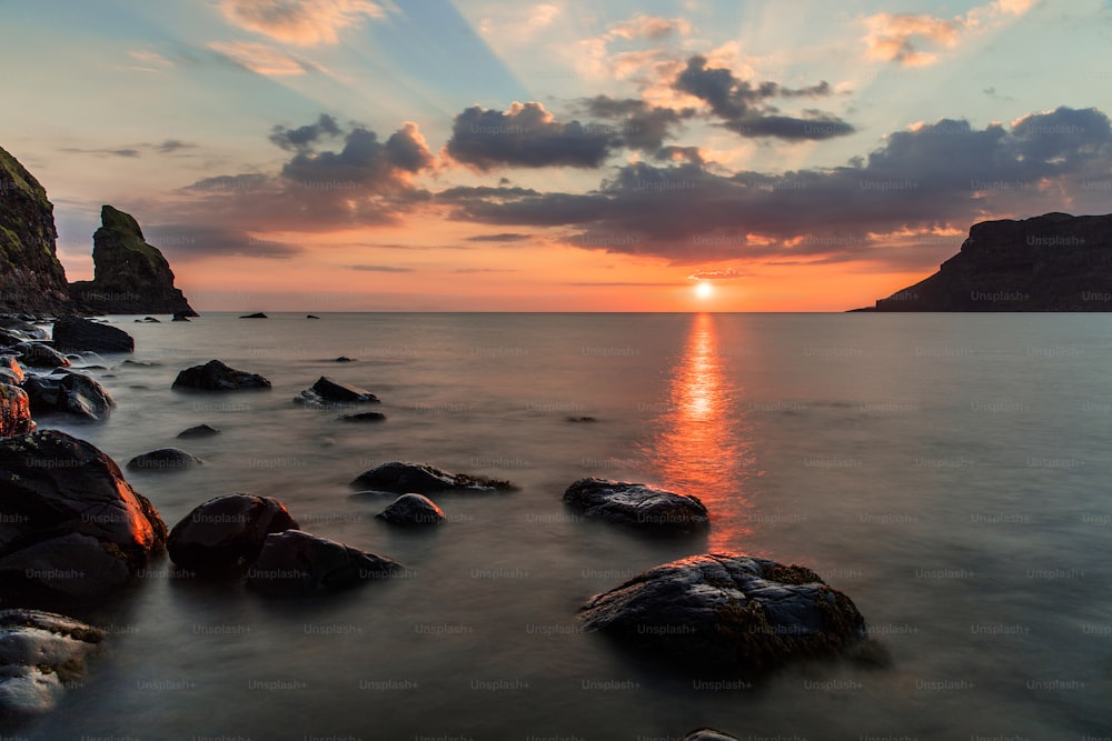 the sun is setting over the ocean with rocks in the foreground