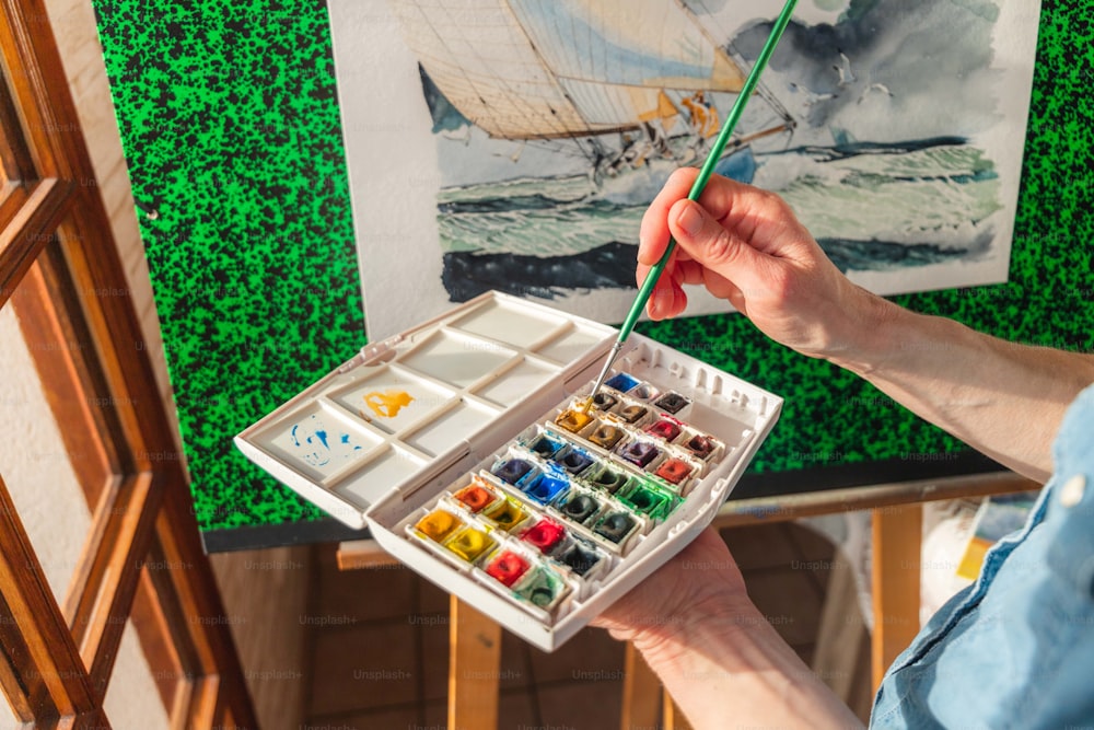 a person holding a paintbrush and a box of watercolors