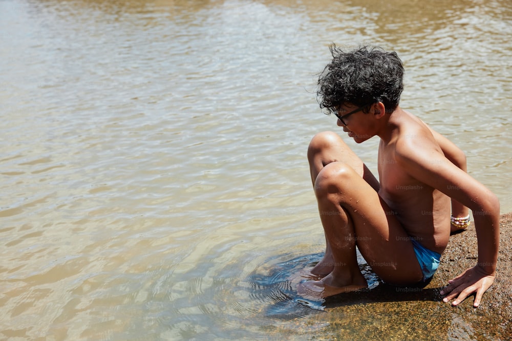 a boy in a blue swimsuit sitting on a rock in the water