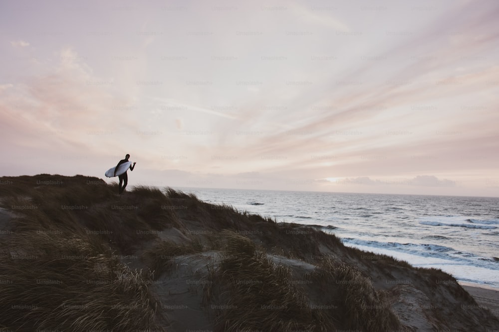 um homem segurando uma prancha de surf em pé no topo de uma praia de areia