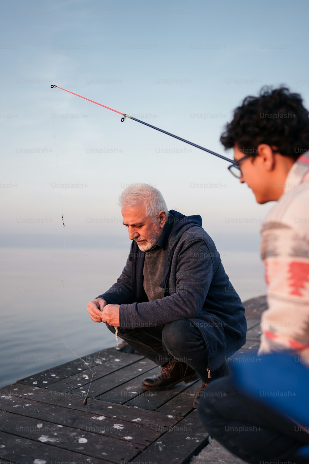 A man sitting on a dock next to another man holding a fishing rod