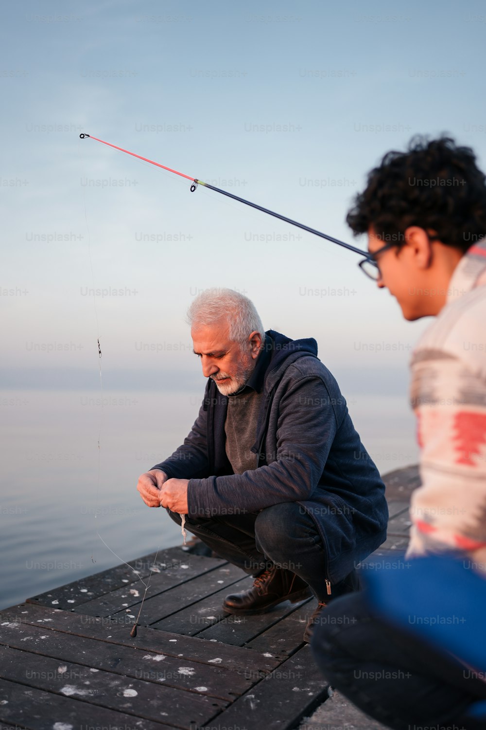 a man sitting on a dock next to another man holding a fishing rod
