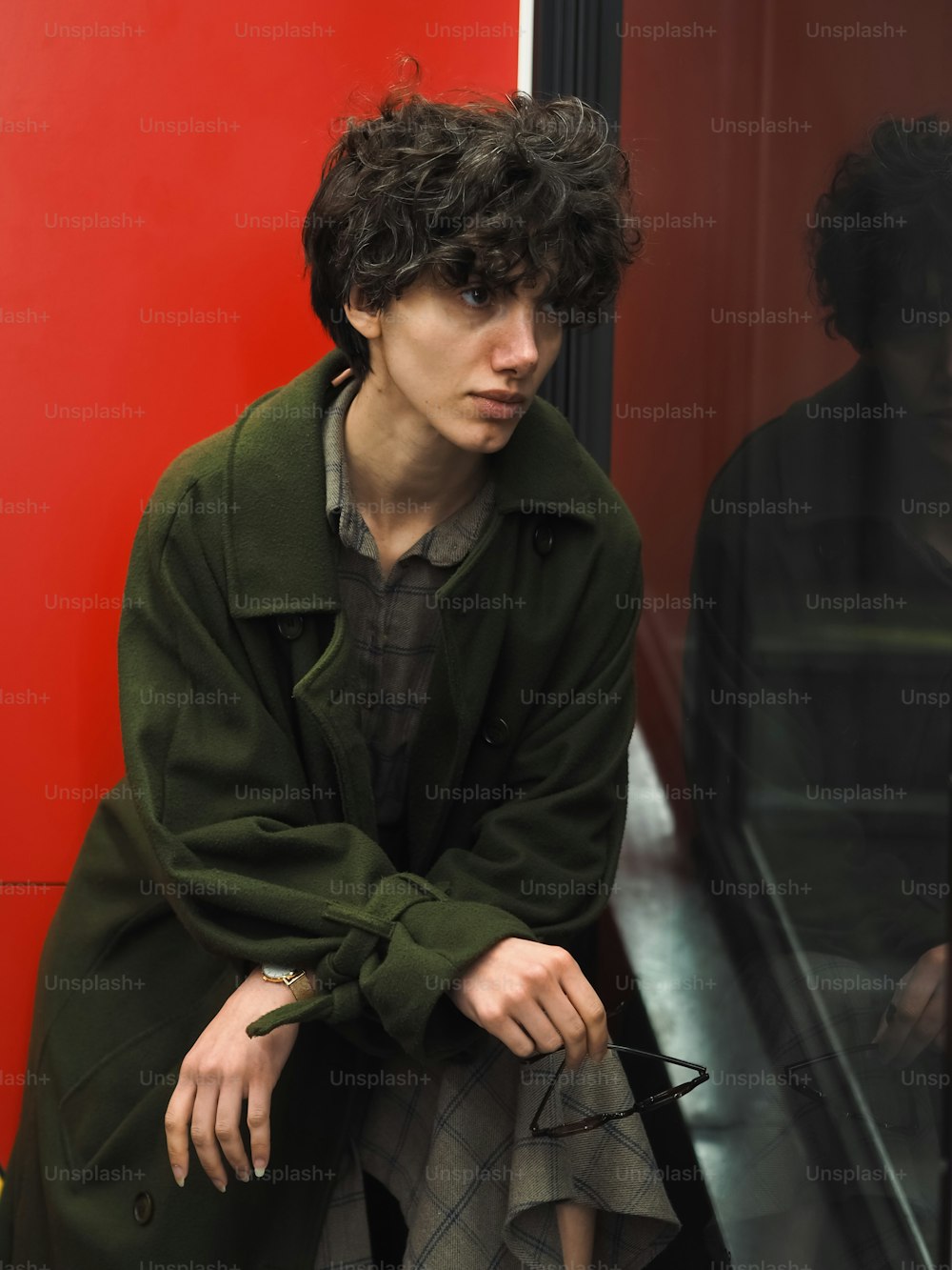 a man in a green coat leaning against a red wall