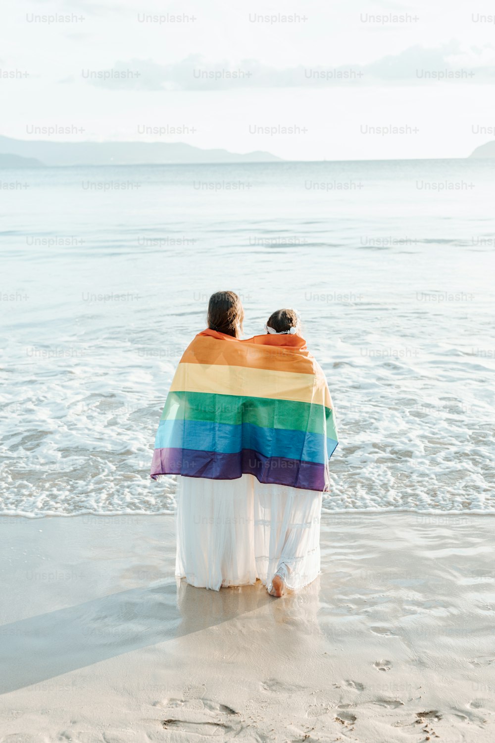 a couple of people sitting on top of a beach under a rainbow blanket