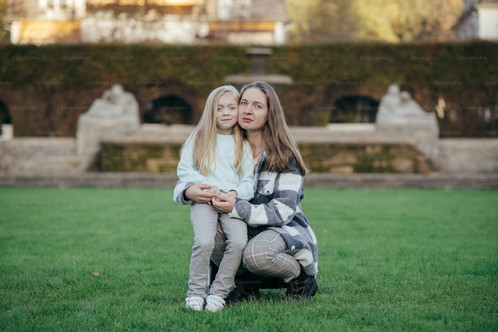 a mother and daughter sitting on the grass in front of a fountain