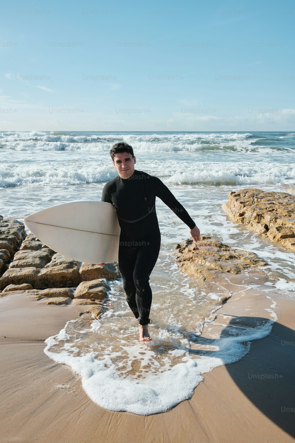 Ein Mann im Neoprenanzug trägt ein Surfbrett am Strand