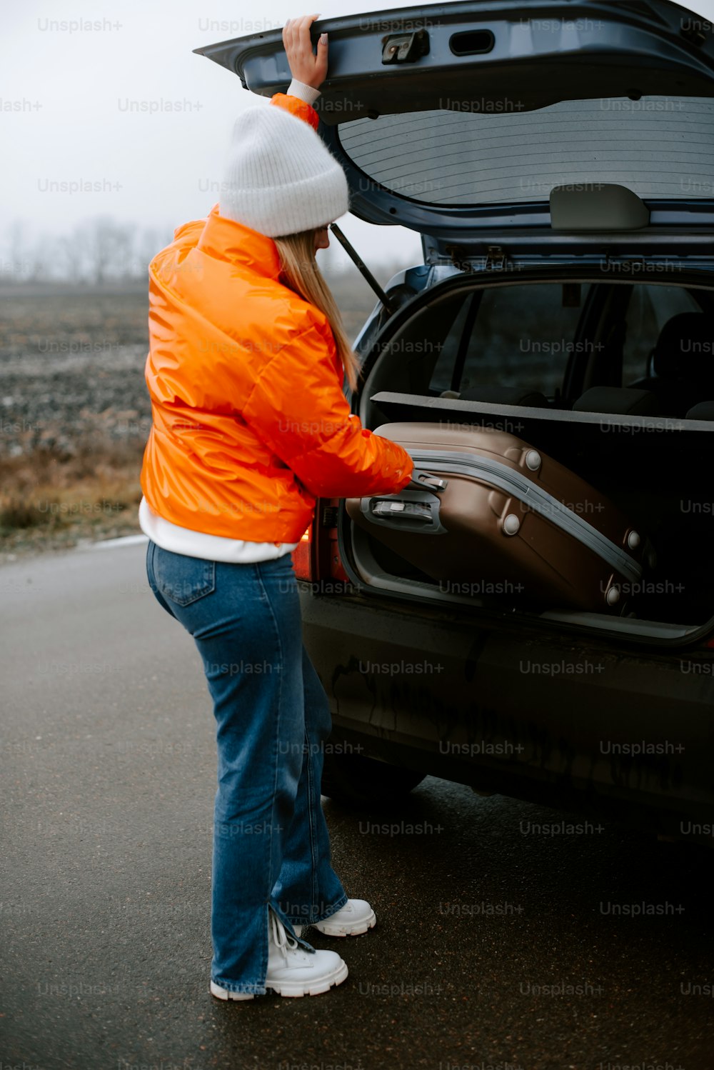a woman in an orange jacket opening the trunk of a car