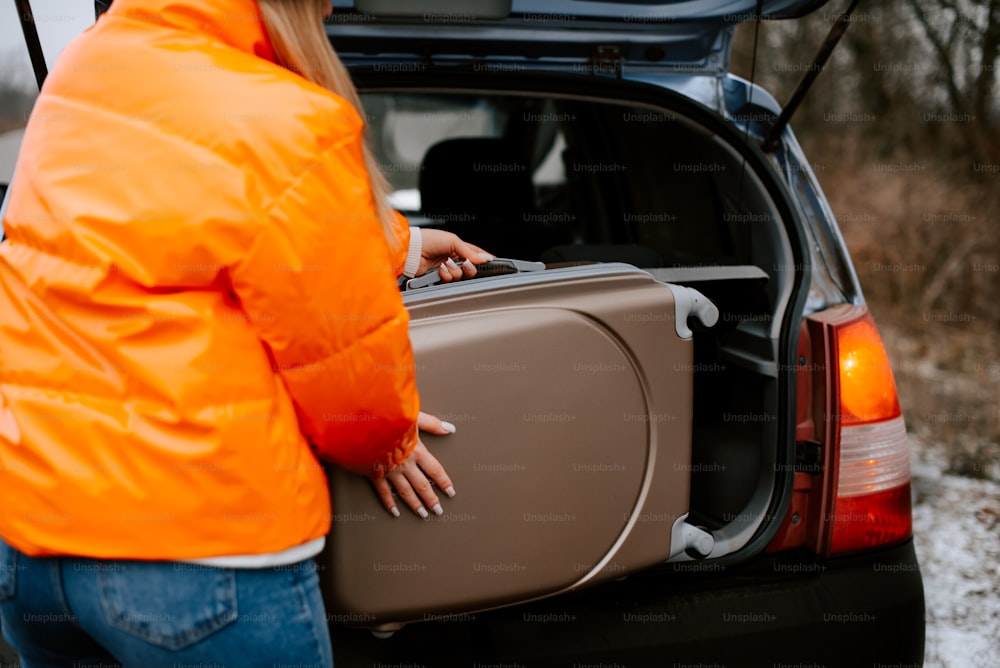 a woman in an orange jacket loading a suitcase into the back of a car
