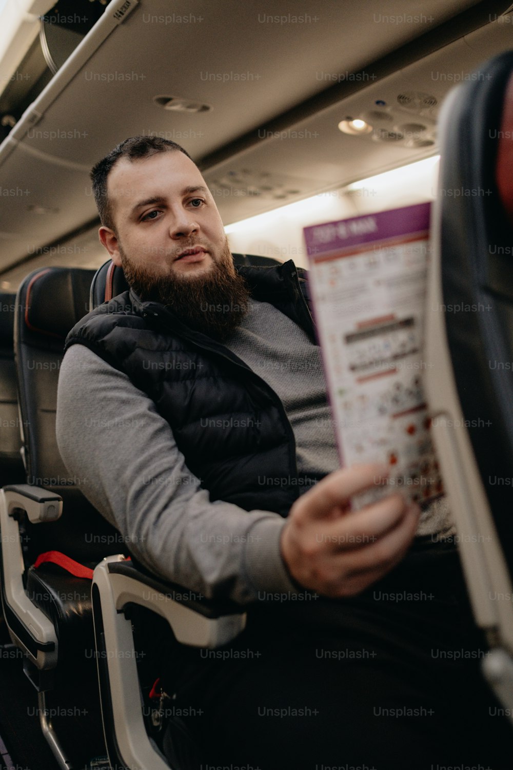a man with a beard sitting on an airplane reading a paper