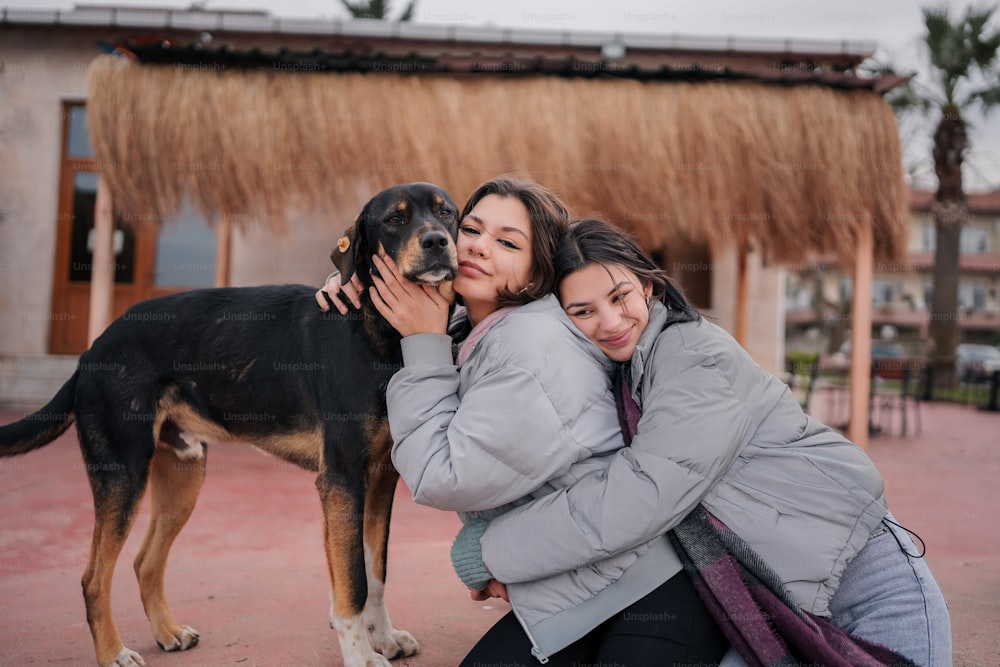 two women hugging a dog in front of a hut