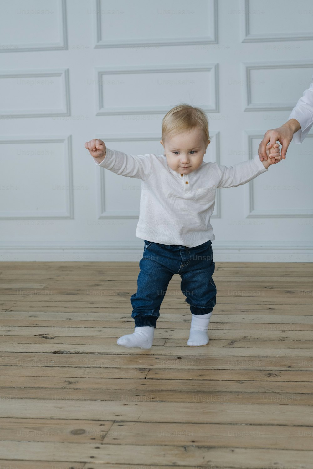 a baby standing on a wooden floor holding the hand of an adult