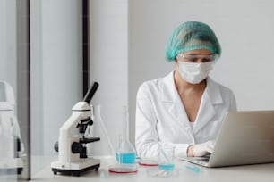 a woman in a white lab coat and mask working on a laptop