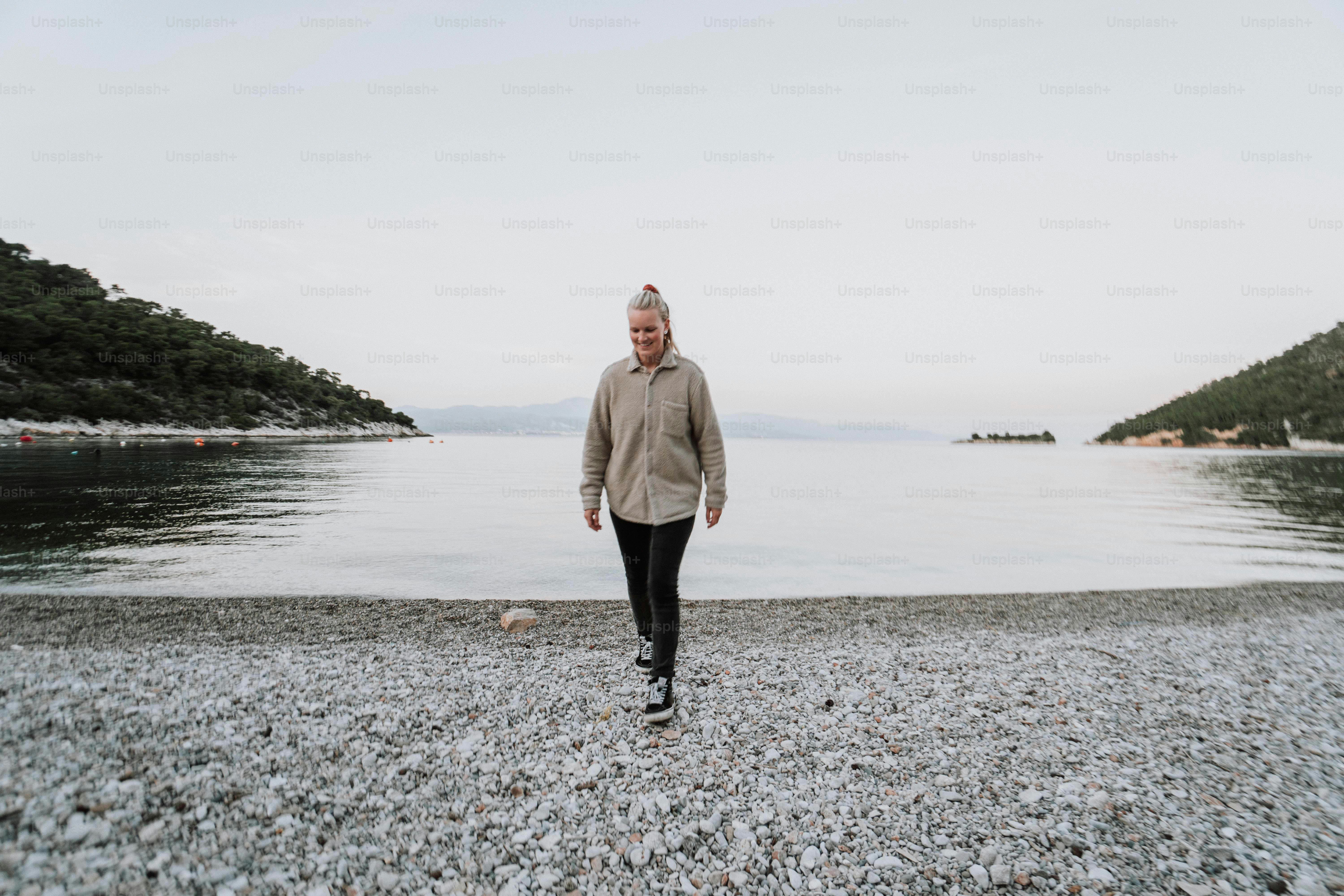 A woman walking on a beach next to a body of water photo – Beach Image ...