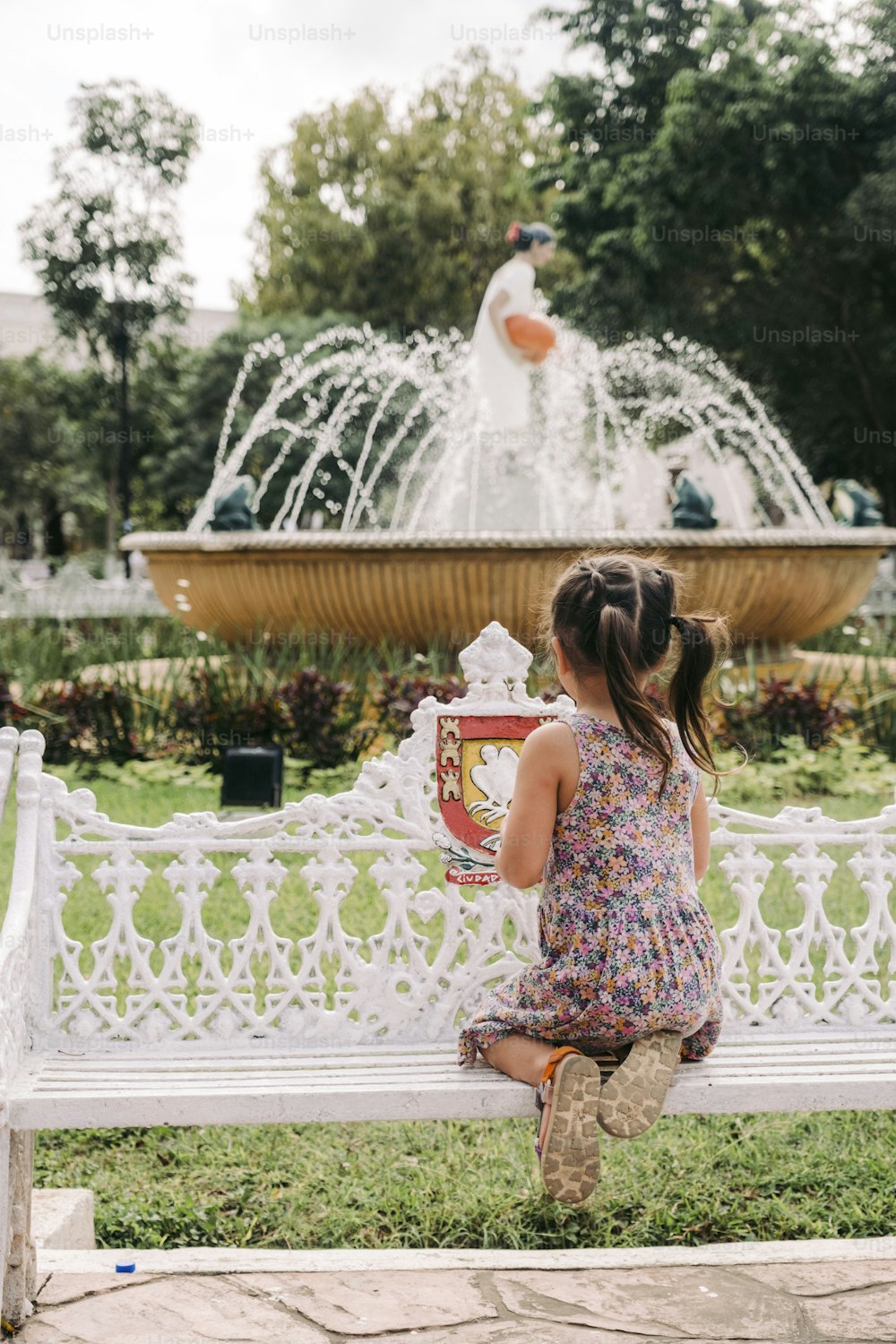 a little girl sitting on a bench in front of a fountain
