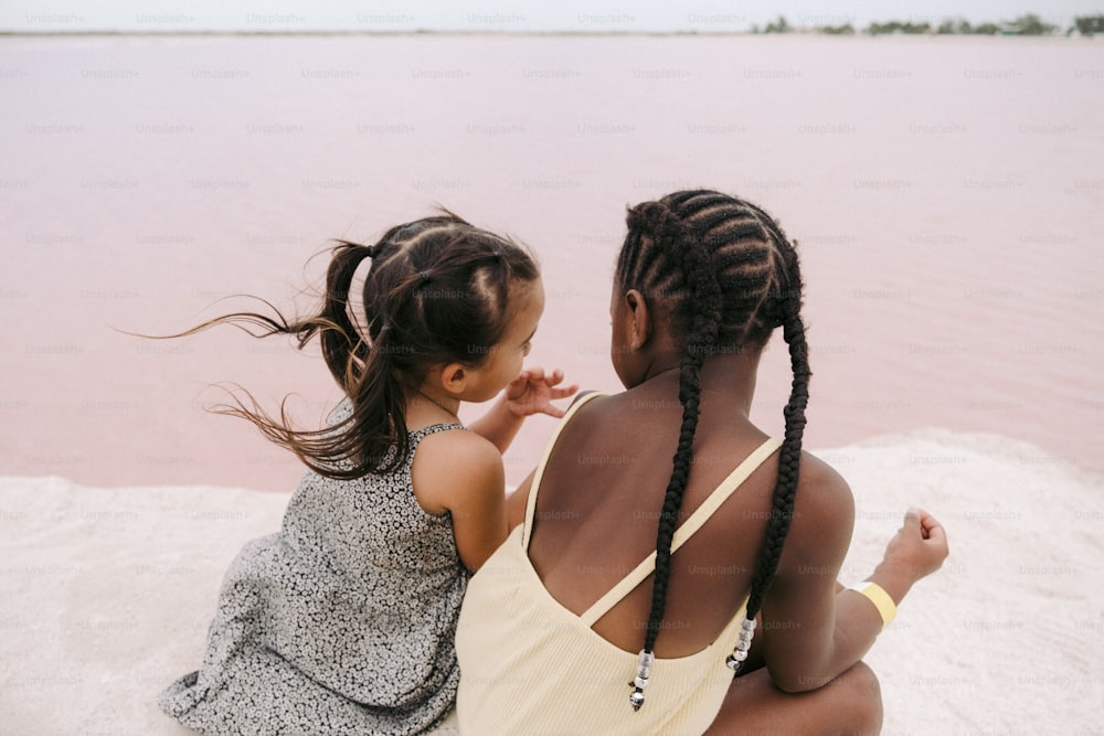 two little girls sitting on the sand near a body of water