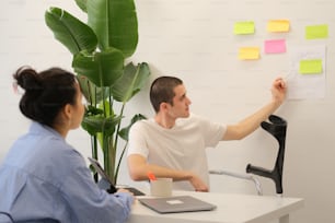 a man and woman sitting at a desk with sticky notes on the wall