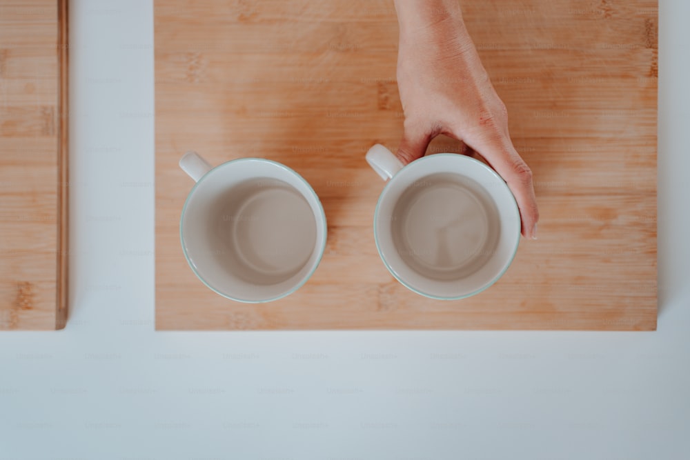 Una persona sosteniendo dos tazas de café encima de una tabla de cortar