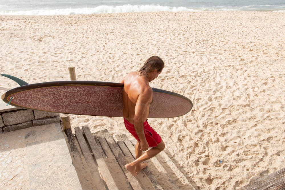 Un hombre sin camisa cargando una tabla de surf en una playa