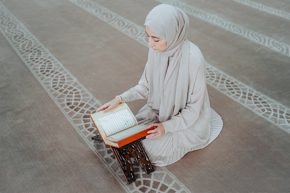 a woman in a hijab sitting on a rug reading a book
