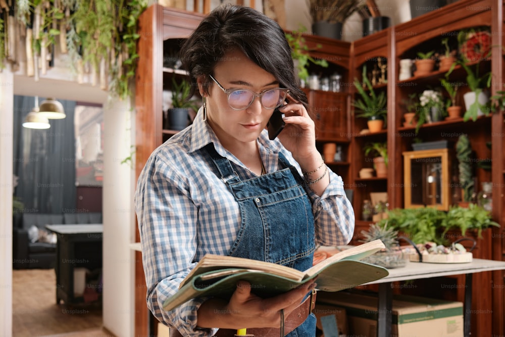 a woman in overalls talking on a cell phone while holding a book